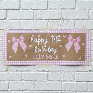 May include: A rectangular sign with a brown center and a pink gingham border. The sign says "happy 18th birthday LILLY GRACE" in white script, with pink bows and hearts. The sign is taped to a white brick wall.