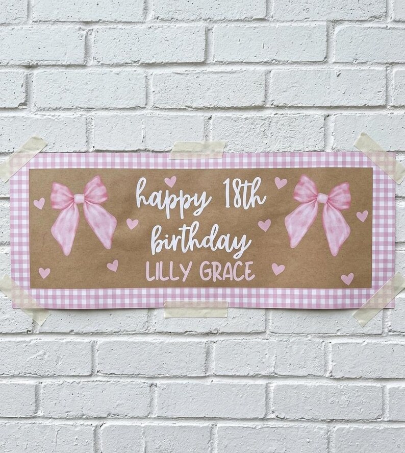 May include: A rectangular sign with a brown center and a pink gingham border. The sign says "happy 18th birthday LILLY GRACE" in white script, with pink bows and hearts. The sign is taped to a white brick wall.