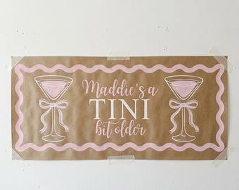 May include: A rectangular sign with the text "Maddie's a TINI bit older" in white and pink. The sign features two martini glasses with pink liquid and bows, and a pink wavy border. The sign is taped to a wall.