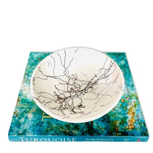 Puede incluir: Un cuenco de cerámica blanca con un diseño abstracto en blanco y negro se encuentra sobre un libro titulado "Turquoise: The World Story of a Fascinating Gemstone" de Joe Darl Lowry y Joe Lowry.