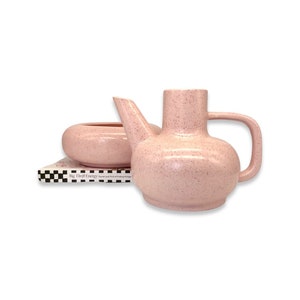 May include: A pink speckled ceramic teapot and a matching bowl. The teapot has a spout and a handle. The bowl is round and has a slight lip. The teapot and bowl are sitting on a book with a black and white checkered cover. The book's title is "Big Third Energy: The Art and Thrift of Finding Vintage."
