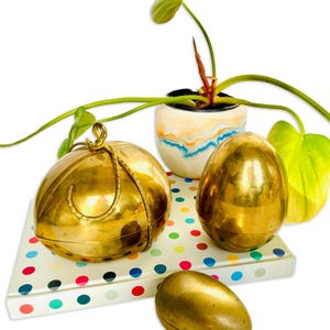 May include: Three gold-colored egg-shaped boxes with a decorative design. The boxes are sitting on a colorful polka dot notebook. A potted plant with green leaves is in the background.