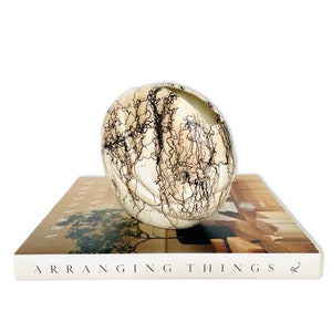 Op de afbeelding: Een witte keramische vaas met zwarte, draaiende lijnen, die op een boek staat met de titel "Dingen ordenen".
