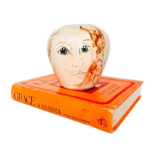 Può includere: Un vaso in ceramica con un ritratto dipinto a mano di una donna con capelli castani e occhi azzurri. Il vaso è appoggiato su un libro intitolato "Grace: A Memoir" di Grace Coddington.