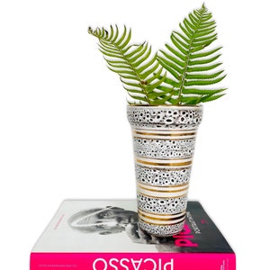 May include: A white ceramic vase with gold stripes and black speckles, filled with green ferns, sits on top of a pink book titled "Picasso: Portrait in Time" by Olivier Widmaier Picasso.
