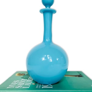 Puede incluir: Una botella de vidrio azul claro con cuerpo redondo y cuello alto y delgado. La botella tiene un tapón redondo azul claro en la parte superior. La botella está sobre un libro verde con el texto "YVES SAINT LAURENT THE SCANDAL COLLECTION 1971".