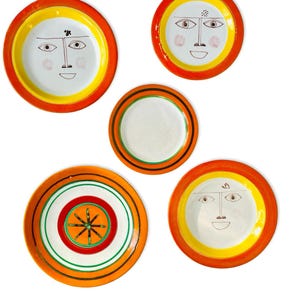 Puede incluir: Colección de cinco platos decorativos de cerámica. Los platos presentan un centro blanco con un borde amarillo y naranja. Algunos platos tienen un diseño de cara, otros patrones geométricos. Adecuado para la decoración de paredes.