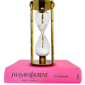 Puede incluir: Un reloj de arena de latón con arena blanca sobre un libro rosa titulado "Yves Saint Laurent Catwalk Haute Couture" de Yale.