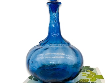 May include: A cobalt blue glass decanter with a spherical body and a long, twisted neck. The decanter has a round stopper and sits atop a book titled "LEAF SUPPLY". The glass has a textured appearance.