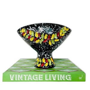 May include: A black ceramic bowl with a white speckled glaze and a floral design in red, yellow, and green. The bowl is on a green and white patterned book titled "Vintage Living".