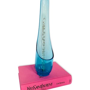 May include: A tall, light blue glass vase with a unique curved shape. The vase is sitting on top of a pink book with the text "Yves Saint Laurent haute couture CATWALK Yale" printed on the cover.