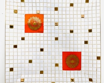 Puede incluir: Una bandeja de cerámica blanca con un patrón de cuadrícula y detalles dorados. Dos azulejos naranjas con detalles dorados están en el centro de la bandeja.