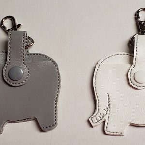 May include: Two leather elephant-shaped keychains. One is gray, the other is white. Each has a silver-tone clasp and a snap closure. The stitching is white. The keychains are designed to hold small items.