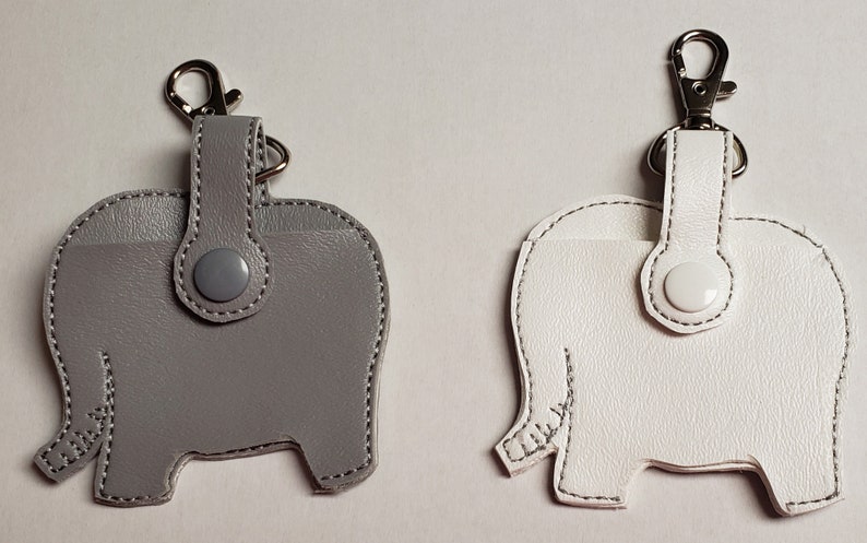 May include: Two leather elephant-shaped keychains. One is gray, the other is white. Each has a silver-tone clasp and a snap closure. The stitching is white. The keychains are designed to hold small items.
