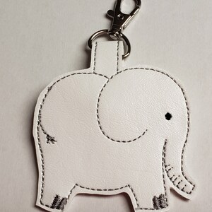 May include: White leatherette elephant keychain with black stitched details. The elephant has a small black eye and a silver-colored clasp. The keychain is a cute accessory.