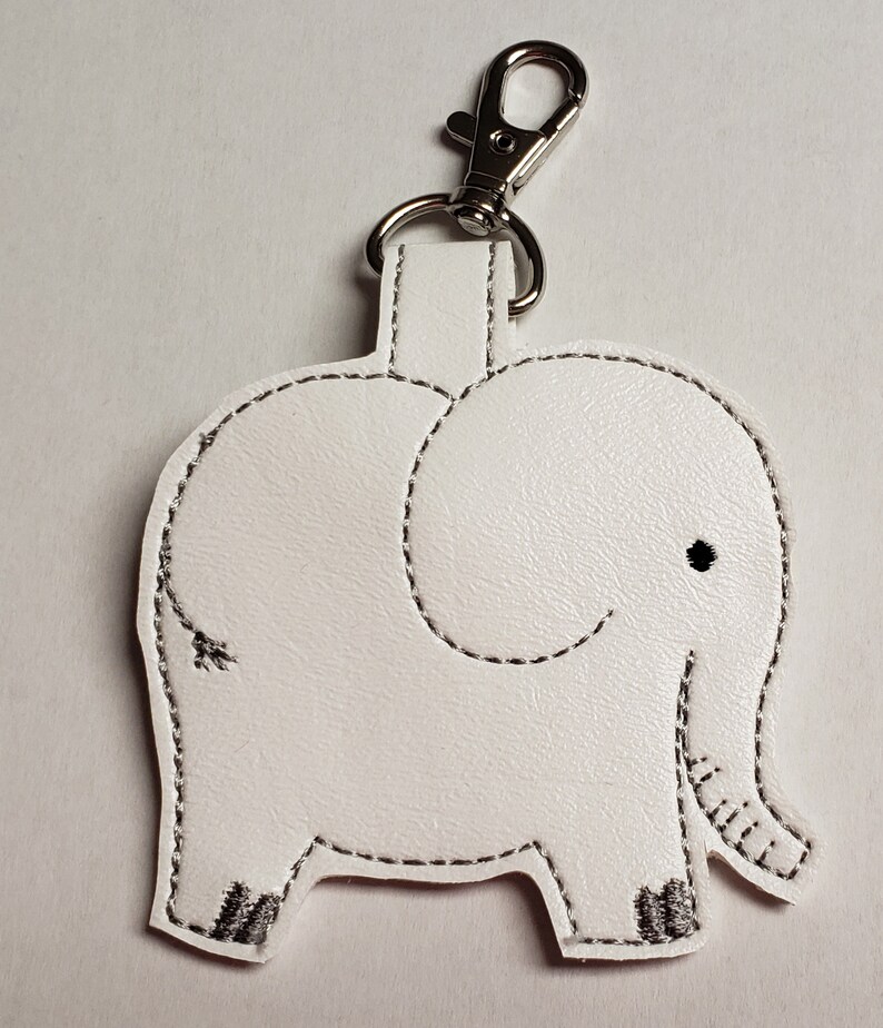 May include: White leatherette elephant keychain with black stitched details. The elephant has a small black eye and a silver-colored clasp. The keychain is a cute accessory.