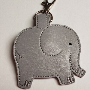 May include: A gray elephant-shaped keychain with a silver clasp. The elephant design features stitched detailing in white thread, including the outline, eye, and foot details. The keychain is made of a textured material.