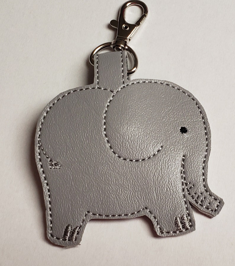 May include: A gray elephant-shaped keychain with a silver clasp. The elephant design features stitched detailing in white thread, including the outline, eye, and foot details. The keychain is made of a textured material.