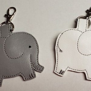 May include: Two elephant-shaped keychains. One is gray, the other is white. Both have black stitched outlines and a silver-colored clasp. The keychains are made of a smooth material and feature a smiling face.