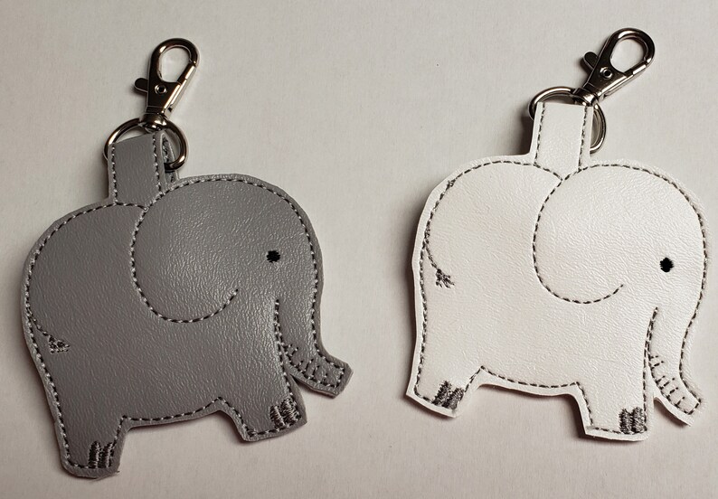 May include: Two elephant-shaped keychains. One is gray, the other is white. Both have black stitched outlines and a silver-colored clasp. The keychains are made of a smooth material and feature a smiling face.