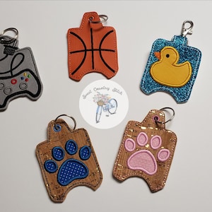 May include: Five different hand sanitizer holders with keychains. The holders are made of different materials and feature various designs, including a basketball, a duck, a paw print, a paw print with polka dots, and a video game controller.
