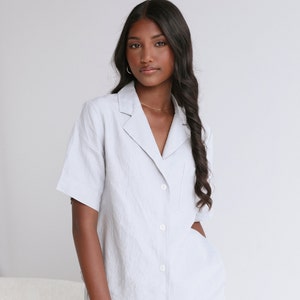 May include: A light blue linen shirt with a collared neckline, short sleeves, and a single button pocket on the left chest. The shirt has a relaxed fit and is button-up.