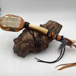 May include: A handcrafted Native American rattle with a light brown wooden handle and a tan leather head. The head features a carved design and is attached to the handle with black leather. The handle is decorated with black leather, beads, and feathers. Tan leather strips hang from the end.