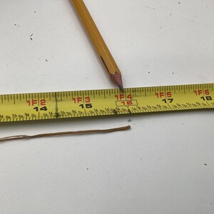 May include: A yellow measuring tape with black markings and a pencil. The tape shows measurements in feet and inches. A brown string lies below the tape. The pencil tip is at the 16-inch mark.