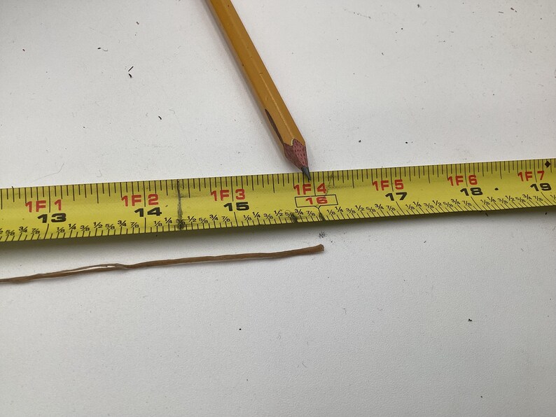 May include: A yellow measuring tape with black markings and a pencil. The tape shows measurements in feet and inches. A brown string lies below the tape. The pencil tip is at the 16-inch mark.