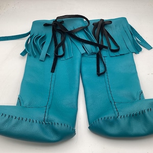 May include: A pair of turquoise leather boots with a moccasin-style toe and fringe detail at the top. The boots have black ties and stitching. The boots are knee-high.