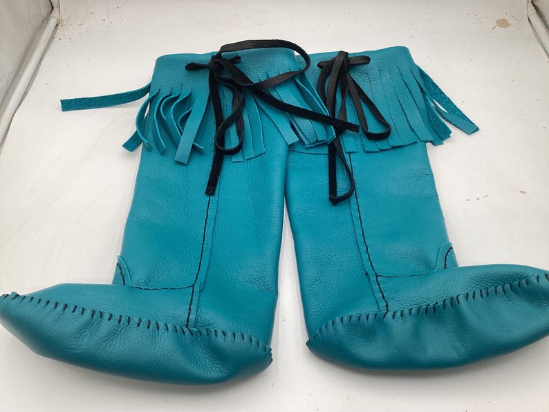 May include: A pair of turquoise leather boots with a moccasin-style toe and fringe detail at the top. The boots have black ties and stitching. The boots are knee-high.