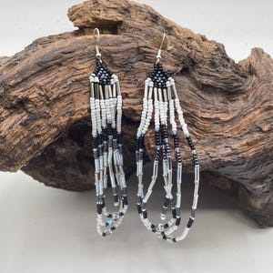 May include: A pair of long, dangling earrings featuring a geometric pattern of small glass beads in black, white, and metallic silver. The design consists of multiple vertical strands of beads suspended from a triangular top section with silver hooks.