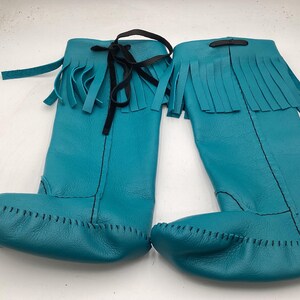 May include: A pair of turquoise leather knee-high boots. The boots feature a fringed top, a black tie closure, and a stitched toe. The boots are made of soft leather and are designed for comfort and style.