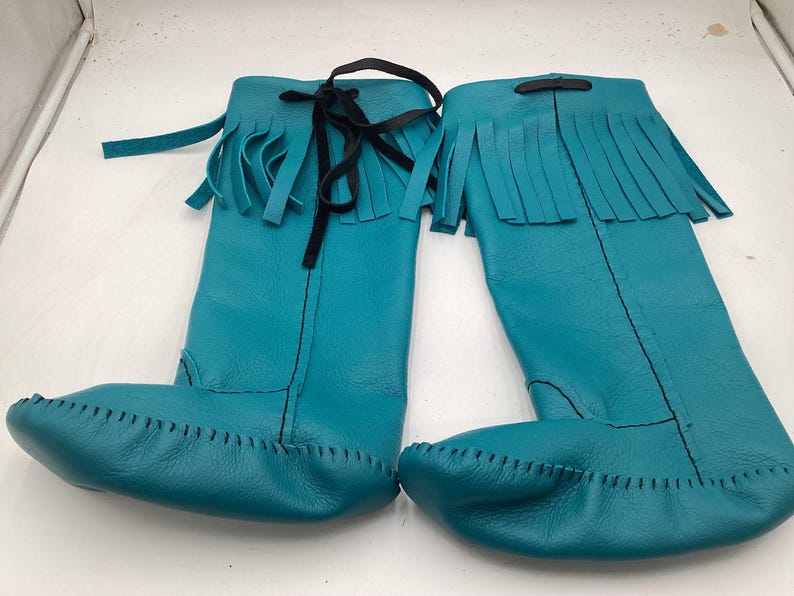 May include: A pair of turquoise leather knee-high boots. The boots feature a fringed top, a black tie closure, and a stitched toe. The boots are made of soft leather and are designed for comfort and style.