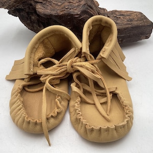 May include: A pair of tan leather moccasins featuring a soft, flexible construction with a gathered toe seam, a fringed collar detail, and thin leather laces tied in a bow across the top of the foot, showcasing a natural, unlined texture.