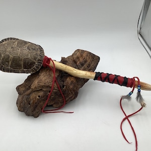 May include: A handcrafted Native American ceremonial rattle. The rattle features a turtle shell head attached to a wooden handle. The handle is wrapped with red and black leather, and adorned with feathers and beads. The rattle rests on a piece of driftwood.