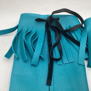 May include: Close-up of turquoise leather moccasin boots with fringe detailing. The boots have a black tie closure and are made of textured leather. The fringe hangs from the top of the boot.