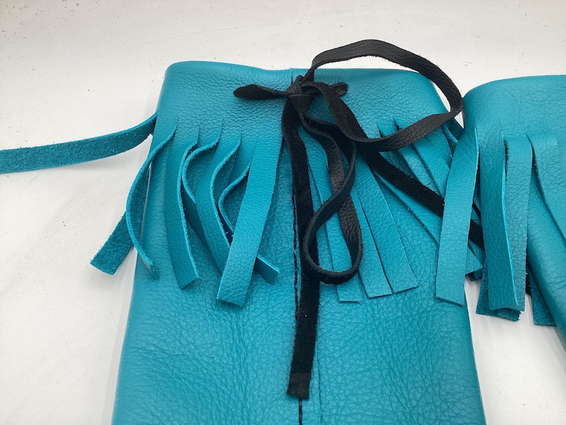 May include: Close-up of turquoise leather moccasin boots with fringe detailing. The boots have a black tie closure and are made of textured leather. The fringe hangs from the top of the boot.