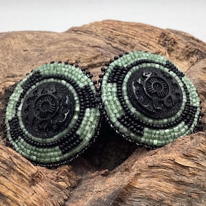 May include: A pair of circular earrings featuring intricate beadwork in alternating bands of sage green and black glass seed beads. At the center of each earring is a black circular component with a cutout sun symbol design and a textured finish.