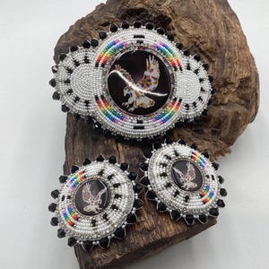 May include: A set of three circular beaded accessories featuring a central black cabochon with a white eagle graphic. The edges are decorated with white seed beads, rainbow-colored accents, and black pointed beads arranged in a radial pattern.