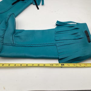 May include: Teal leather moccasin boots with fringe detail. The boots are approximately 12 inches long, with black stitching and a black leather strap. The boots are laid out on a white surface with a yellow measuring tape.