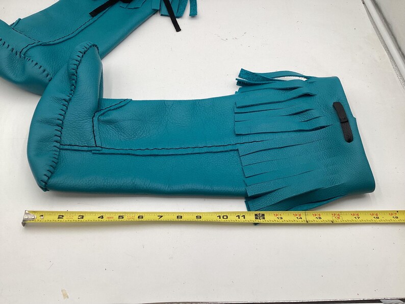 May include: Teal leather moccasin boots with fringe detail. The boots are approximately 12 inches long, with black stitching and a black leather strap. The boots are laid out on a white surface with a yellow measuring tape.