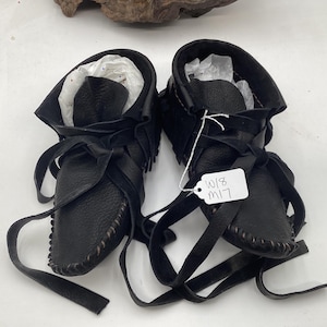 May include: A pair of black leather moccasin-style boots with long, wraparound ties. The boots have a soft, textured appearance and are lined with white paper. The stitching is visible along the seams. A tag with the text "W18 M17" is attached.