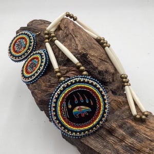 May include: A necklace and matching pair of earrings featuring circular beaded medallions with a bear paw and bear silhouette design in rainbow colors on a black background, accented with white hair pipe beads and metallic bronze spacer beads.