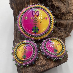 May include: A set of three circular beaded accessories featuring a vibrant gradient of pink and yellow. Each piece displays a central floral graphic with green, purple, and orange accents, surrounded by concentric rows of yellow and pink seed beads.