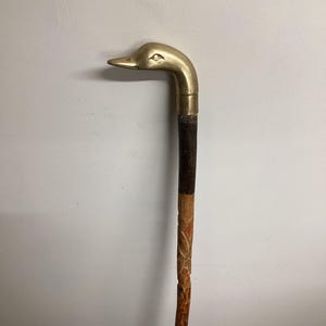 May include: A vintage walking cane with a brass duck head handle. The cane has a dark brown, textured grip and a carved wooden shaft with a natural finish. The duck's eye is detailed.