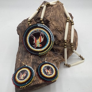 May include: A necklace and earring set featuring circular medallions with beaded borders in black, white, yellow, and red. The center of each piece displays a graphic of an eagle in flight. The necklace includes long white bone-like beads and small metallic spacers.