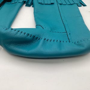 May include: Close-up of turquoise leather moccasins with fringe detail. The moccasins are hand-stitched with black thread and have a soft, textured appearance. The design features a classic, traditional style.