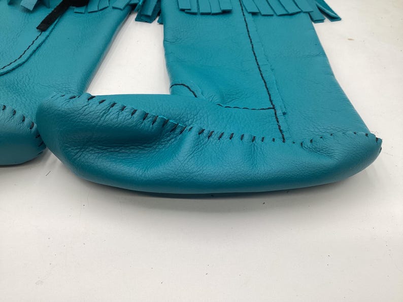 May include: Close-up of turquoise leather moccasins with fringe detail. The moccasins are hand-stitched with black thread and have a soft, textured appearance. The design features a classic, traditional style.