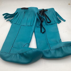 May include: A pair of turquoise leather moccasin boots with fringe detail at the top. The boots have a knee-high design with black ties and stitching. The soles are stitched with a contrasting thread.